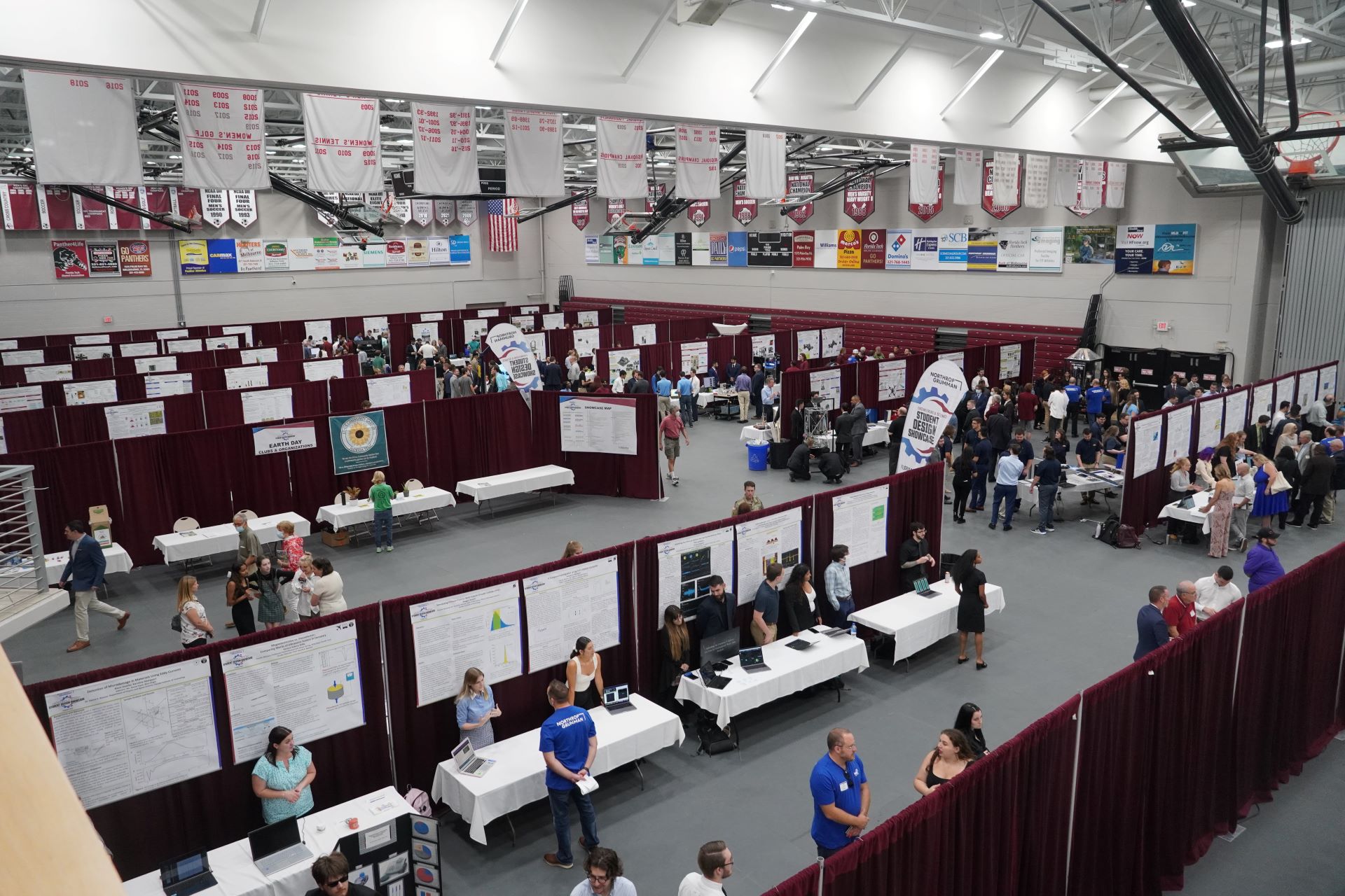 An overhead view of a large event at Florida Tech featuring numerous booths and posters set up in rows. Attendees are walking around, engaging with exhibitors, and reading posters. Banners and advertisements are visible on the walls